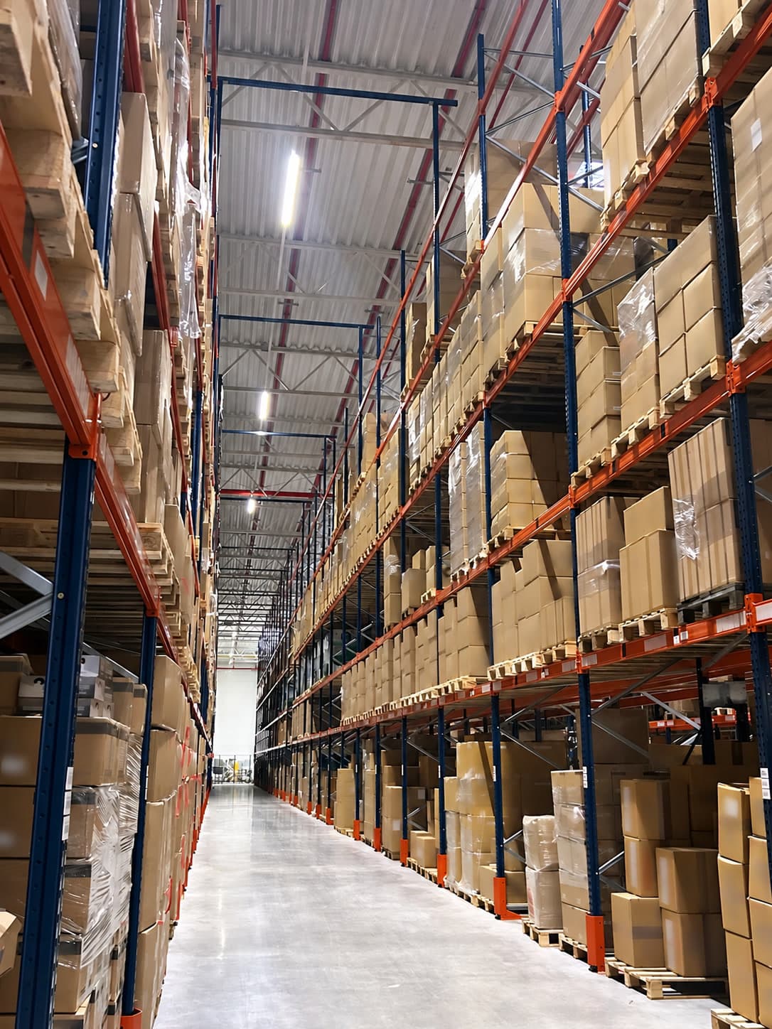 An aisle in our UK warehouse: high pallet racking either side, stacked with cartons, product labels blurred.